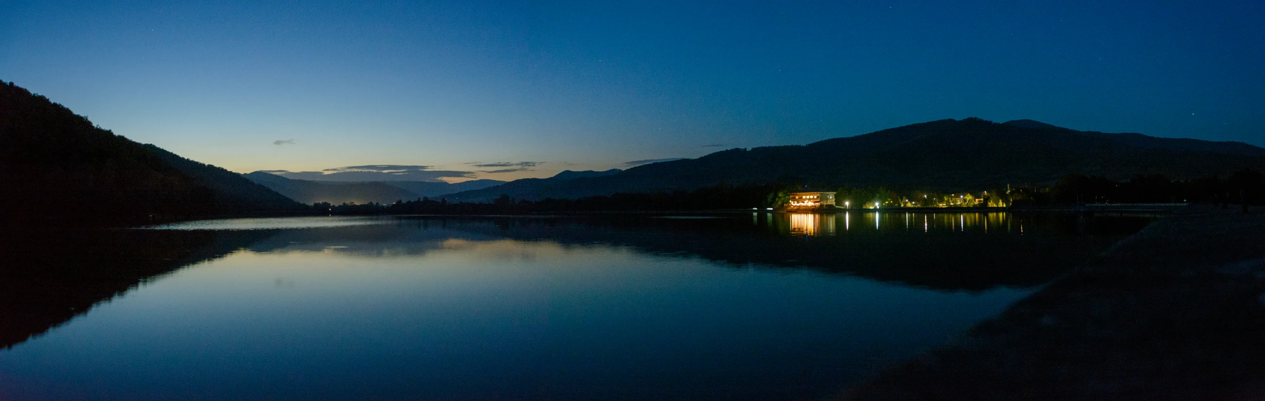 Široká panoráma jazera, kopcov a odrazov svetiel počas modrej hodiny